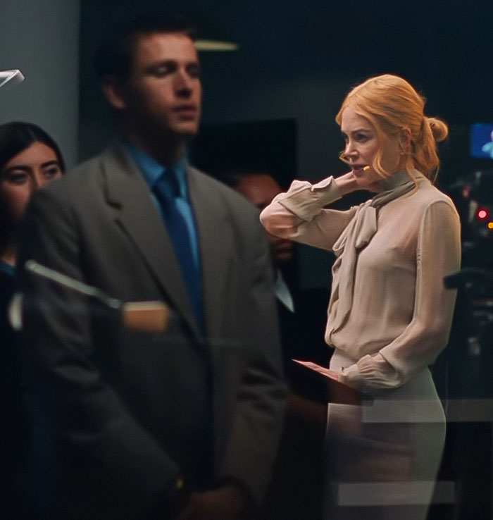 Nicole Kidman, 57, in a serious scene wearing light blouse and skirt, captured in a dimly lit room with blurred figures around.