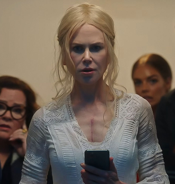 Nicole Kidman, 57, wearing a white lace top, holding a phone, with concerned people in the background.