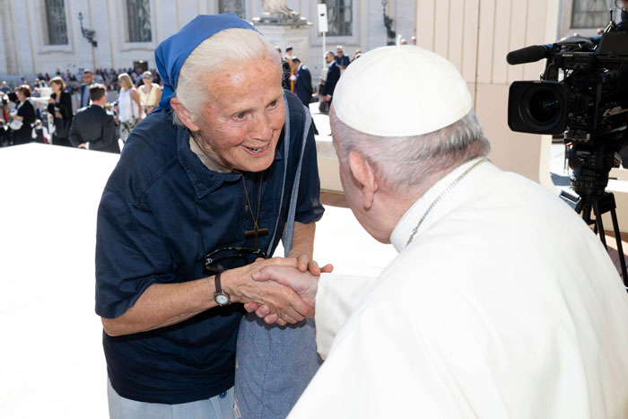 Nun smiling and shaking hands with the Pope, standing outdoors surrounded by people, camera capturing the moment.