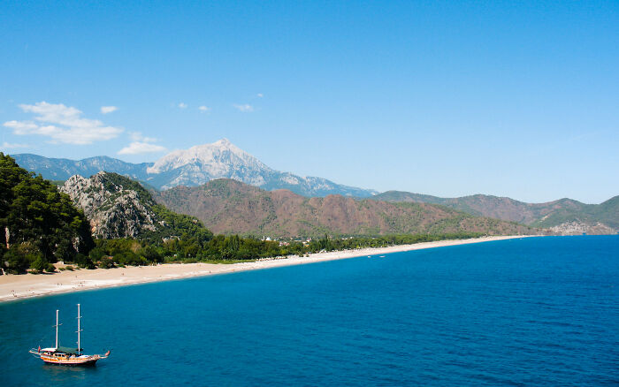 Coastal vista featuring a sandy beach, turquoise sea, lush hills, and a small sailboat under a clear blue sky.