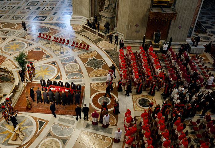 Pope Francis' open casket in Vatican viewed by attendees in a grand hall with intricate floor designs.