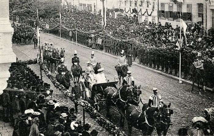 Black and white photo showing life in Europe 100 years ago with a horse-drawn carriage and crowds lining the street.