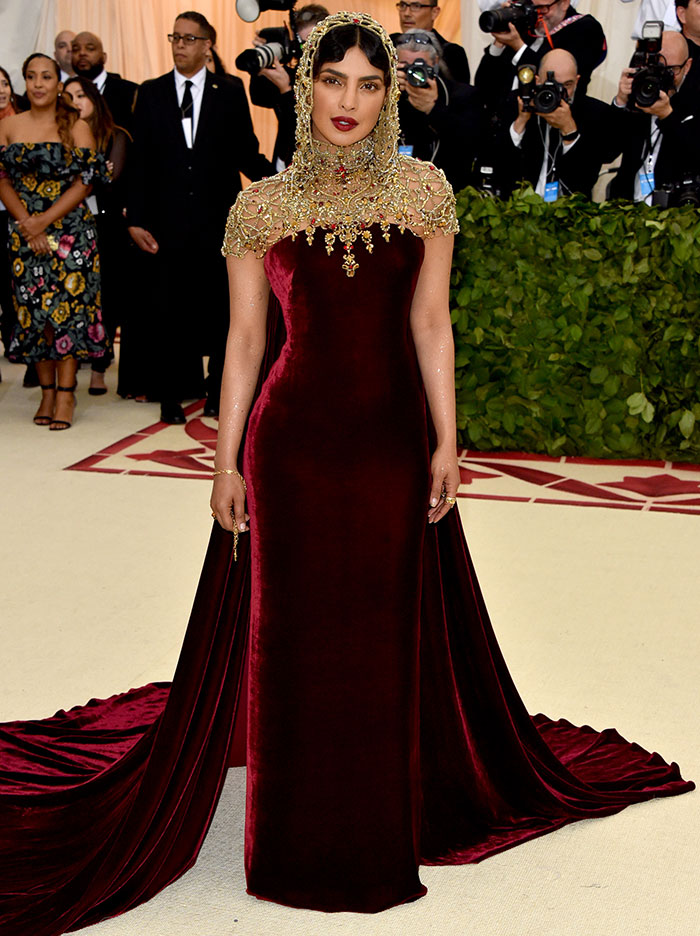 Celebrity in a burgundy velvet gown with gold embellishments on the red carpet; photographers capturing the moment.