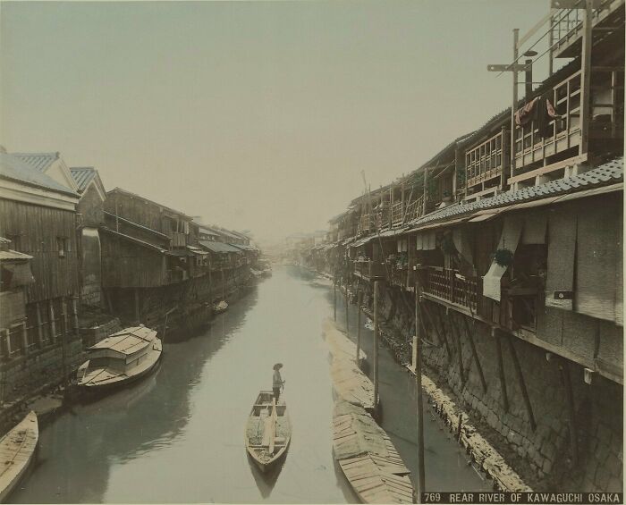 A vintage scene of Japan in the 1800s showing a river with wooden houses and a person navigating a boat in Kawaguchi, Osaka.