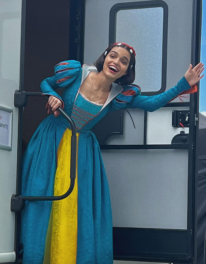 Woman dressed as Snow White stepping out of a trailer, wearing a blue and yellow dress, smiling widely.