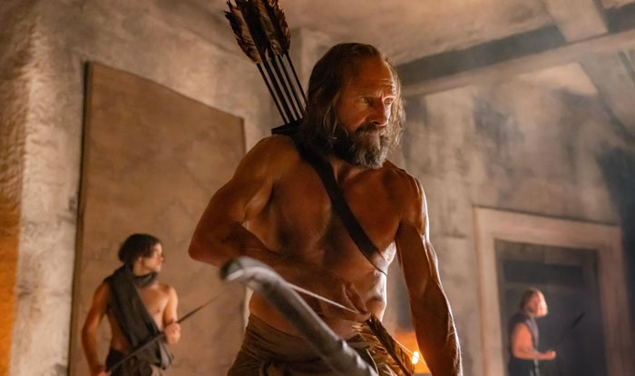 A man with a muscular physique, wearing a shoulder strap and holding a bow, stands in a rustic setting. A man with a muscular physique, wearing a shoulder strap and holding a bow, stands in a rustic setting.