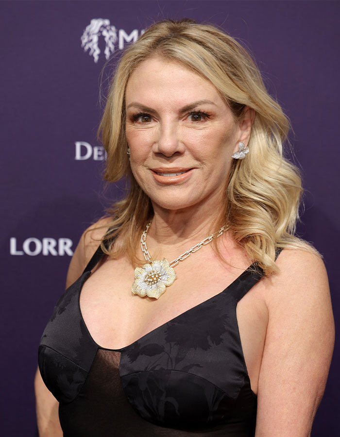 Woman in black dress smiling, with blonde hair and floral necklace, on red carpet.