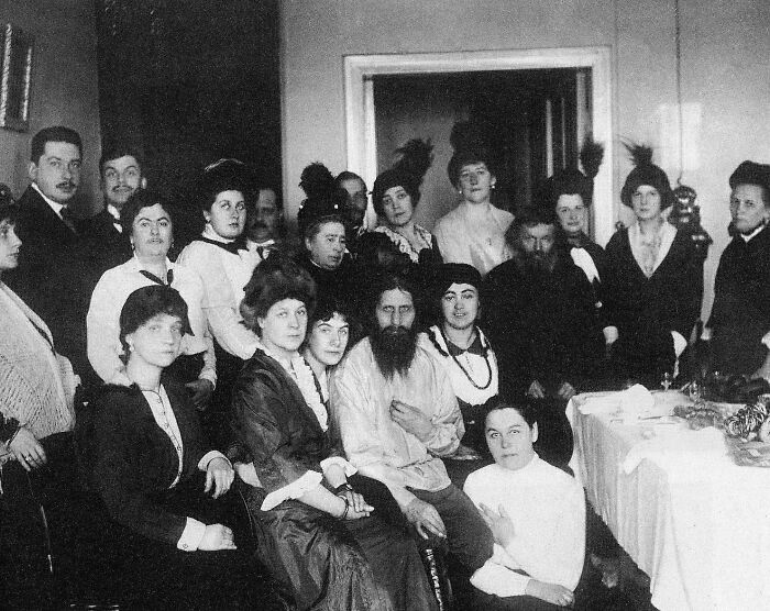 Group of famous historical figures and women gathered in a room, dressed in early 20th century clothing, rare historical photo.