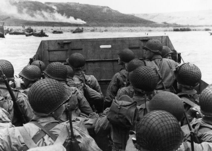 WWII soldiers in helmets on a landing craft approaching a beach, smoke in the distance.