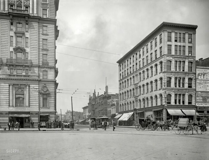 Historic street scene in America from 100 years ago showing buildings, horse-drawn carriages, and pedestrians.