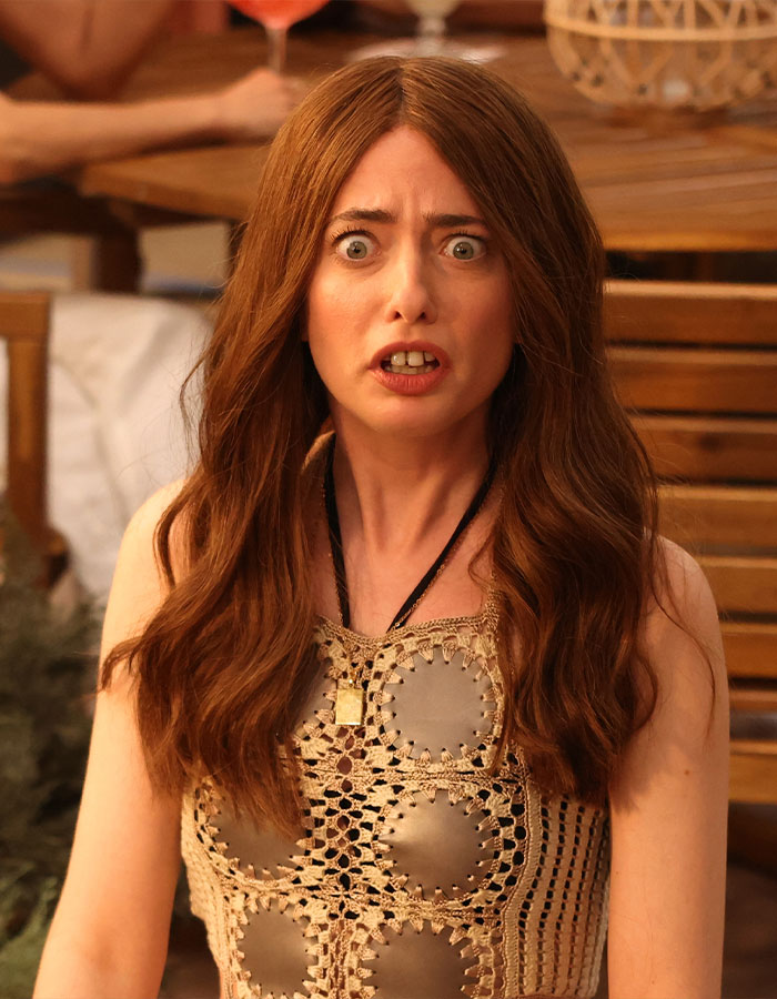 Actress in a surprised expression, seated outdoors, wearing a crochet top at a wooden table, 'White Lotus' star scene.