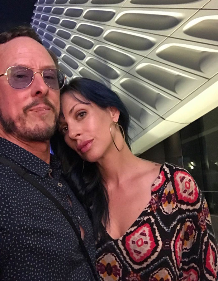 A couple posing in front of a modern architectural background, related to Weezer bassist and lawyer costs discussion.