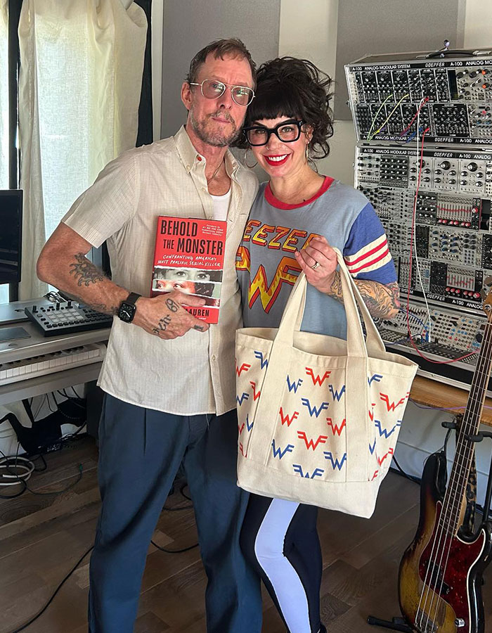 Weezer bassist in a studio, holding a book, next to a woman with a Weezer tote and shirt, near a music synthesizer.