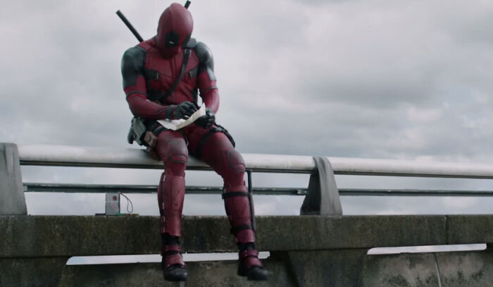 Deadpool in full costume sitting on a ledge, writing on a notepad showcasing things that look awesome in movies.