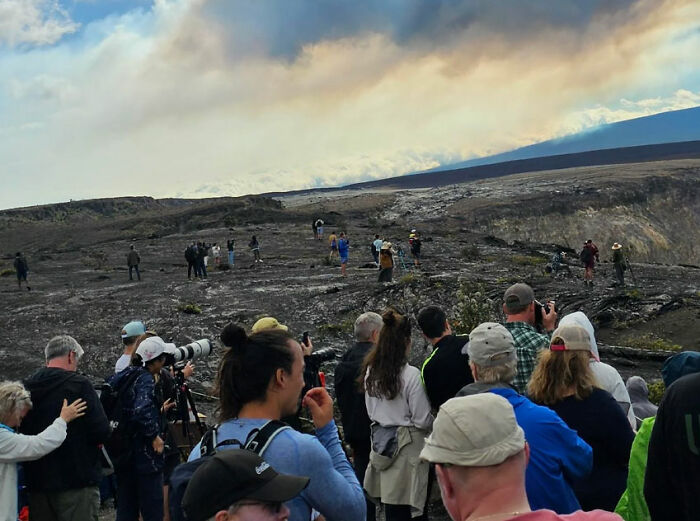 Crowd with cameras at volcanic landscape, some ignoring safety, highlighting lack of self-awareness.