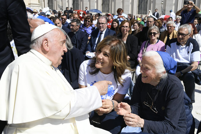 Pope Francis talking to seated nun and woman in crowd, engaging warmly in a public gathering.