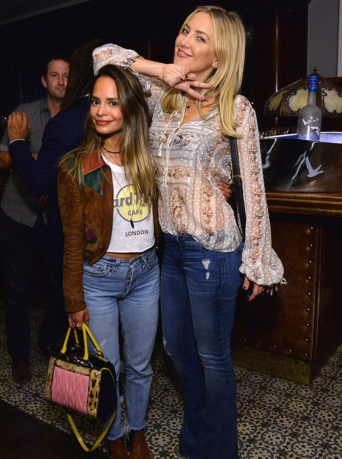 Two women pose in stylish casual outfits at an unglamorous red carpet behind-the-scenes event.
