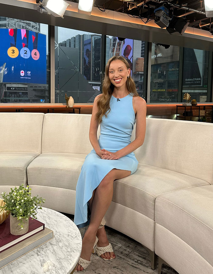 TV reporter in a blue dress sitting on a couch in a studio set.