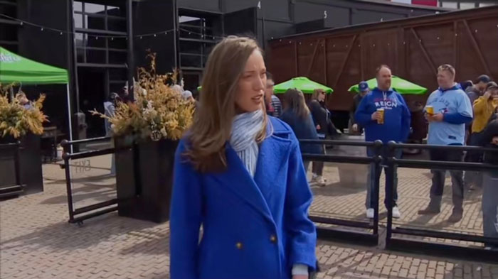 TV reporter in a blue coat appears shocked in a public outdoor setting with people in the background.