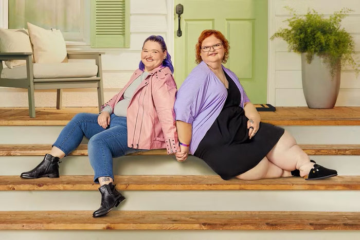 Two women seated on porch steps, smiling, one in a pink jacket and the other in purple, showcasing weight transformation.