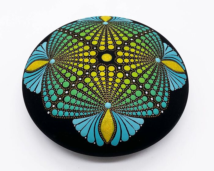 Colorful mandala art on a smooth black stone showcasing intricate dot patterns in yellow, green, and blue.