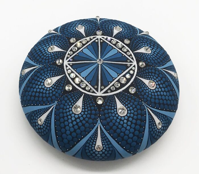 Intricately painted mandala on a smooth stone featuring hypnotic blue and white patterns with decorative gems.
