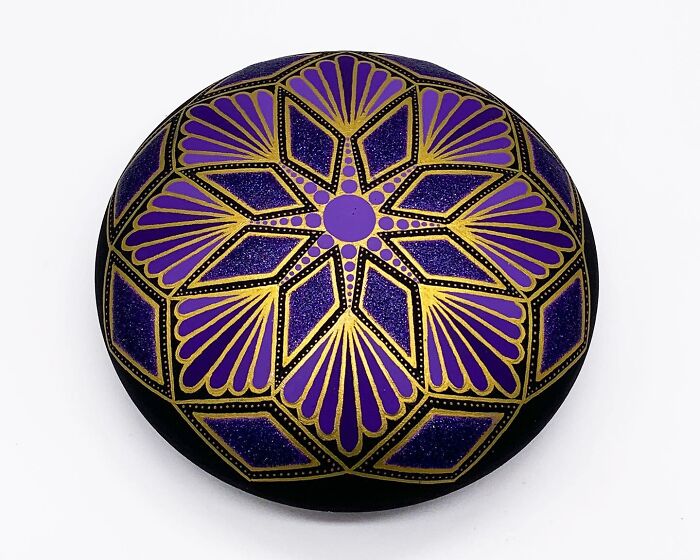 Intricate mandala artwork painted on a stone with purple and gold colors, showcasing beautiful geometric patterns.