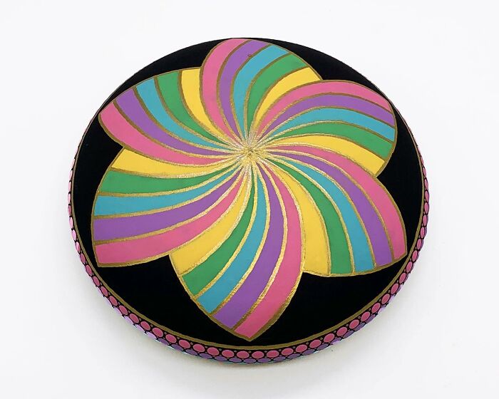 Colorful mandala art on a stone, featuring swirling petal design in pink, yellow, and blue hues with gold accents.