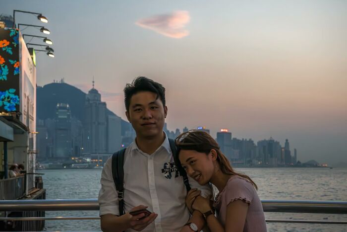 A couple stands by the waterfront at sunset with a heart-shaped cloud above, showcasing a perfectly timed picture.