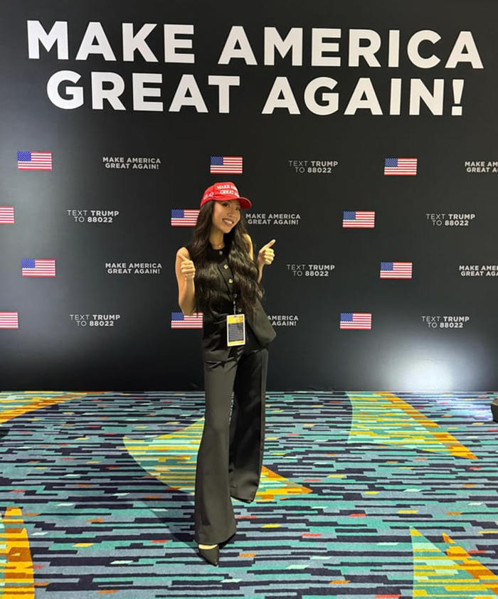Influencer at a MAGA event, wearing a red cap, standing in front of a "Make America Great Again" sign.