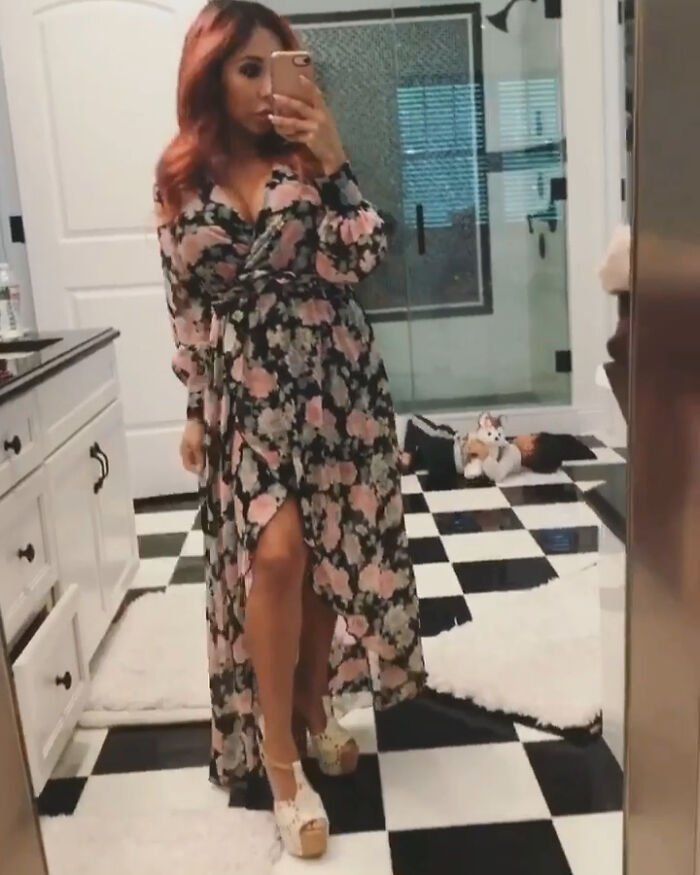 Woman in floral dress taking a mirror selfie in a bathroom with a child lying on a checkered floor, celebrity encounters theme.
