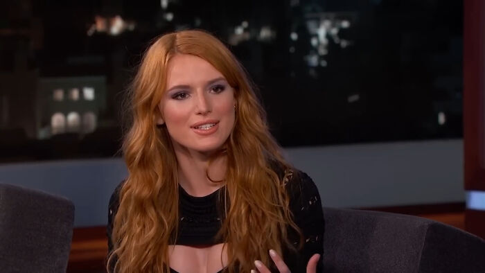 Young woman with long red hair speaking during a nighttime celebrity interview on a talk show set.