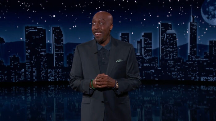 Comedian standing on stage with city skyline backdrop at night, relating stories about celebrity home break-ins.