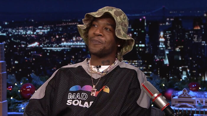 Celebrity guest wearing a camouflage bucket hat and black Mad Solar shirt during a nighttime talk show interview.