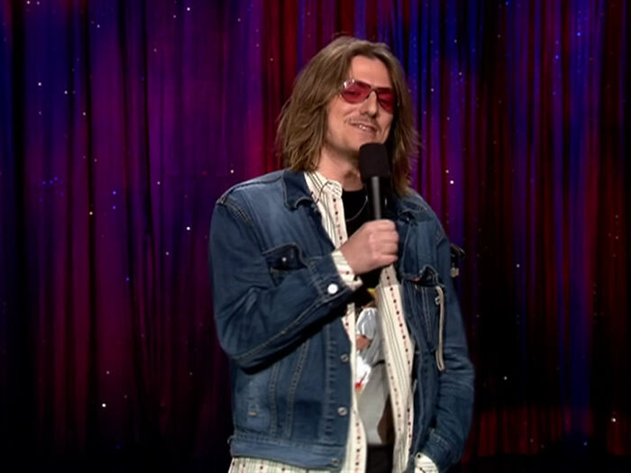 Person on stage with microphone, wearing red sunglasses and denim jacket, in front of red curtain, associated with April Fools.