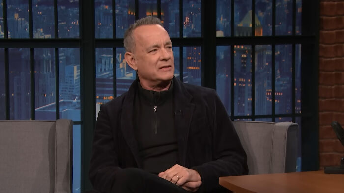 Man in a black jacket sitting on a late night talk show set discussing celebrity home break-ins and security concerns.