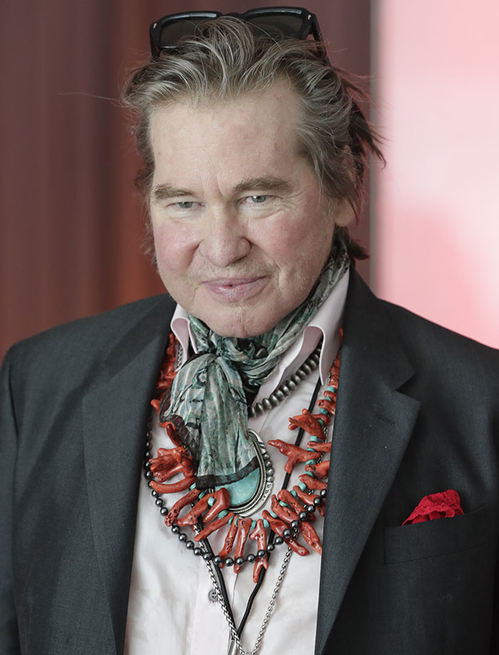 Man in a suit with a colorful necklace and scarf, wearing sunglasses on head and smiling gently in a formal setting.