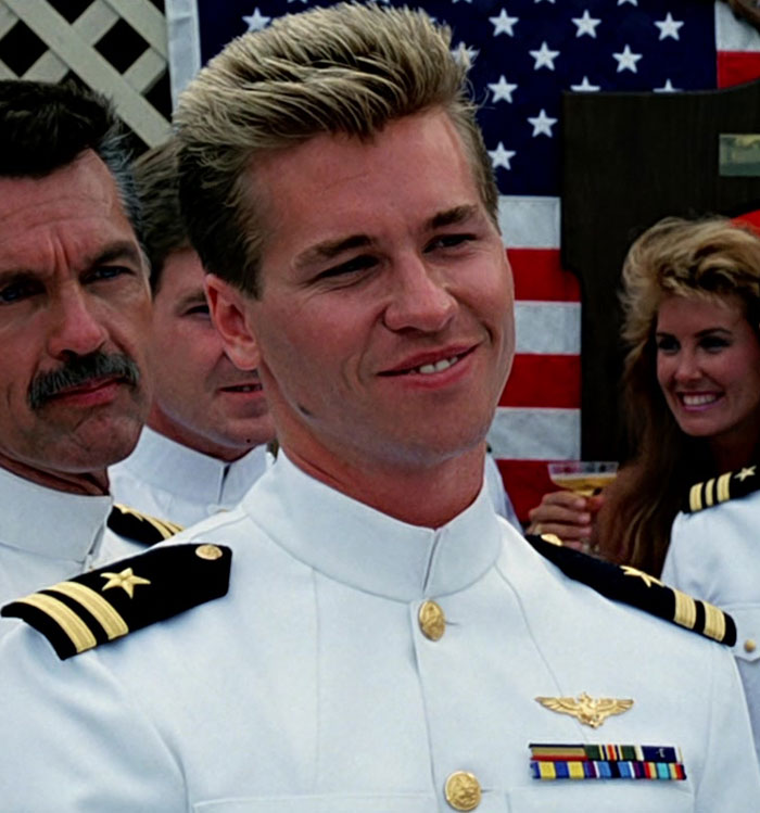 Val Kilmer in a naval uniform, smiling, with an American flag backdrop.