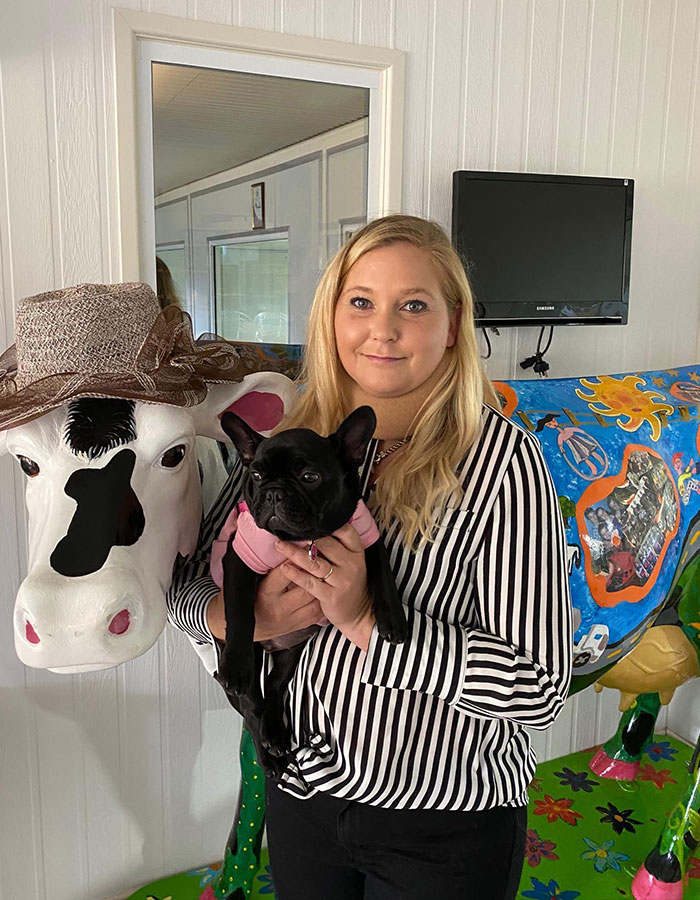 Woman in striped shirt holding a black dog, standing next to a decorated cow sculpture.