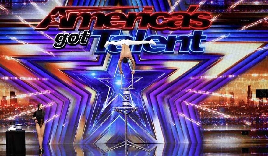 Acrobat performing on stage, balancing on chairs, with a large America's Got Talent logo in the background.