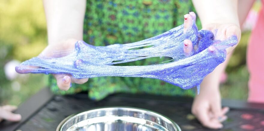 Hands stretching glittery blue slime, a popular viral trend, over a metal bowl outdoors.