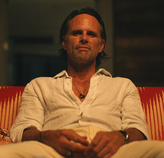 53YO Walton Goggins in photoshoot, seated on a patterned red couch, wearing a white shirt, exuding confidence.