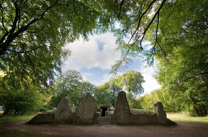 Ancient man-made stone structure surrounded by greenery and tall trees under a blue sky with clouds.