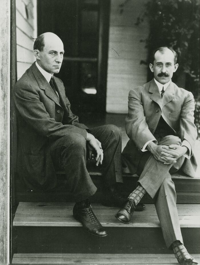 Two famous historical figures sitting on wooden steps outside a building, wearing early 20th-century suits.