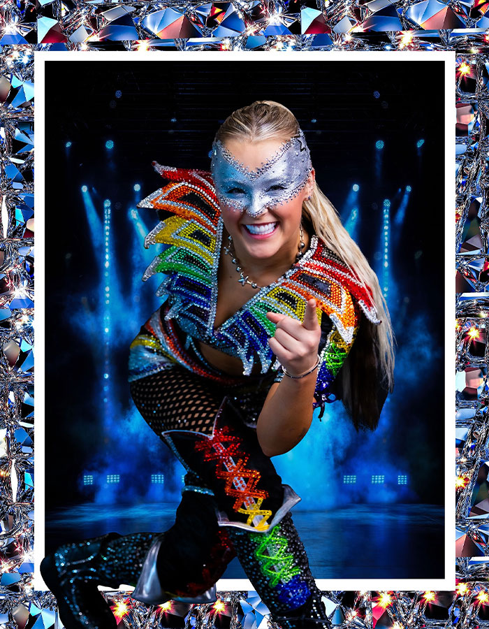 JoJo Siwa in a vibrant rainbow costume and mask, smiling energetically on stage background.
