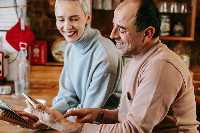 A woman and man smiling at a smartphone in a cozy kitchen setting, discussing family dynamics and relationships. A woman and man smiling at a smartphone in a cozy kitchen setting, discussing family dynamics and relationships.