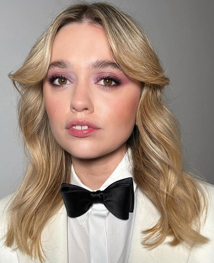 Blonde woman in a white outfit with a bow tie, associated with The White Lotus, highlighting her natural teeth.
