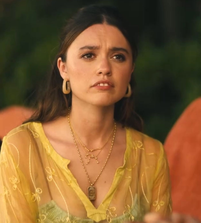 Woman in a yellow blouse with layered necklaces, reacting emotionally, outdoors.