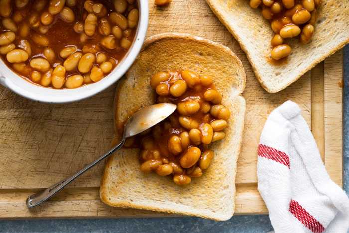Beans on toast served with a side of baked beans and spoon, laid out on a wooden cutting board.