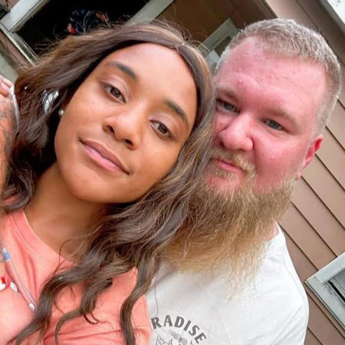 A Black woman and a white man sitting closely, expressions relaxed, outside a building.